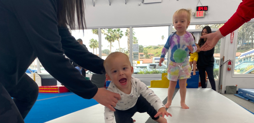 A smiling baby crawling, being helped as a toddler watches