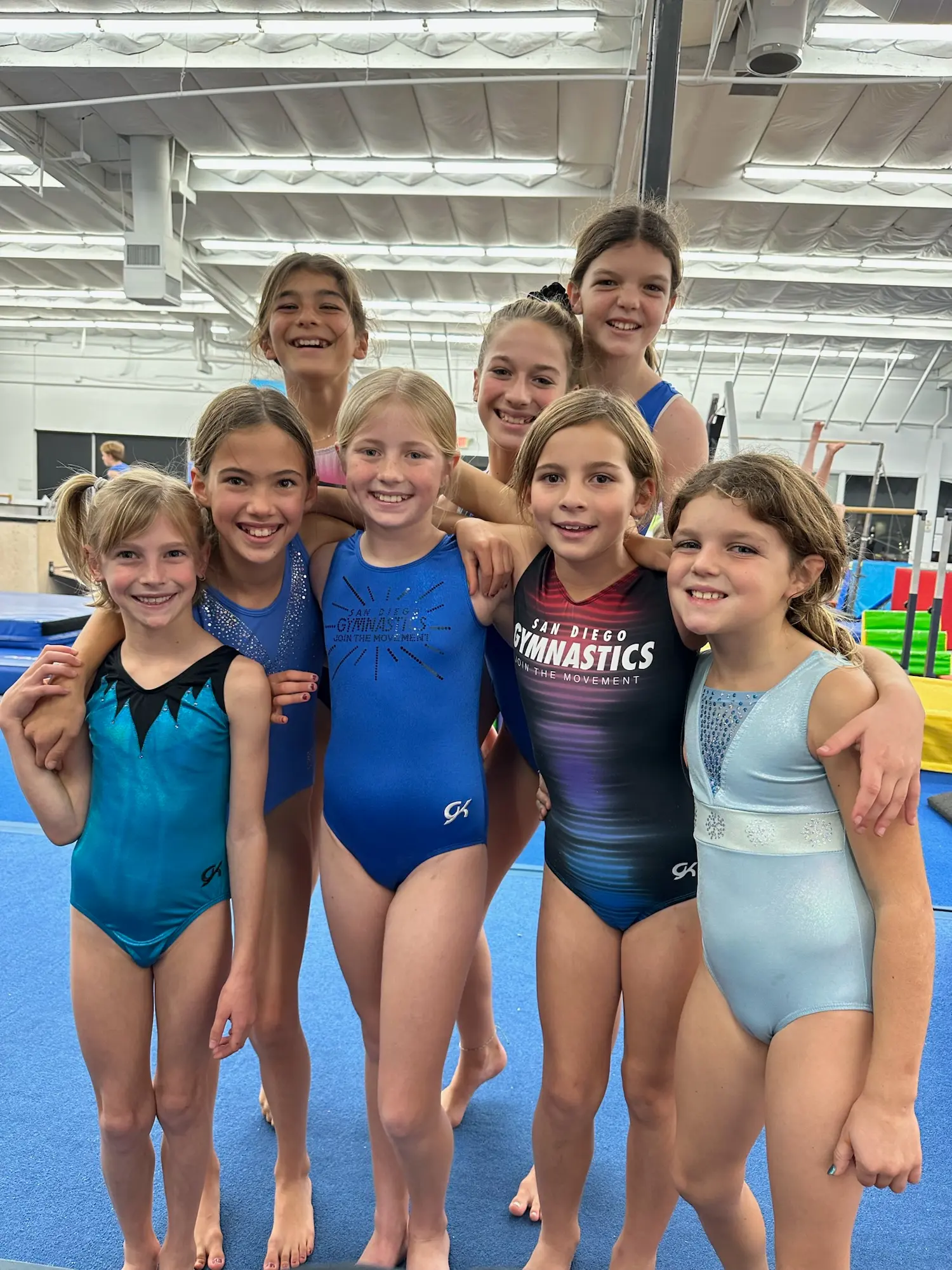 Eight young girls in colorful leotards stand together on a blue mat at a gymnastics gym. They are smiling and embracing, conveying friendship and joy.