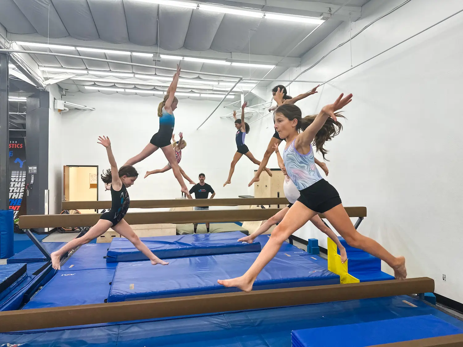 Children in colorful leotards practice leaps on blue mats in a gymnastics facility, exuding energy and focus. A coach observes in the background.